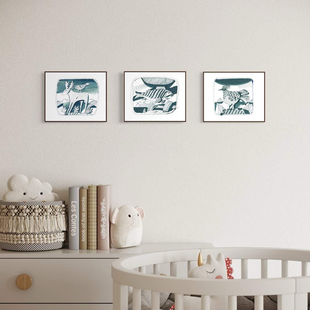 Three framed botanical art prints in a muted teal and white style are displayed on a light beige wall above a nursery dresser. The scene includes a white crib with a plush toy, a woven basket with books and a cloud-shaped pillow, and decorative nursery props.